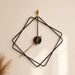 A minimalist wall clock with a black wireframe diamond design and gold-toned hands, mounted on a warm beige wall with a dried grass accent in the lower left corner.