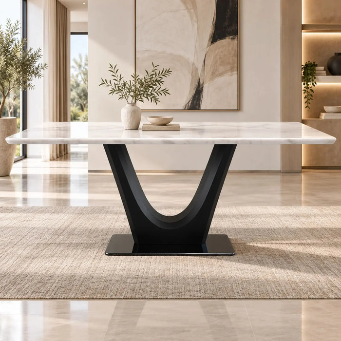 A modern rectangular dining table with a white marble top and a sculptural black U-shaped pedestal base, styled in a bright, contemporary dining room with a beige rug and abstract wall art.