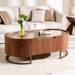 An oval wooden coffee table with a glass top and brass metal legs, styled in a modern living room setting in front of a beige sofa.