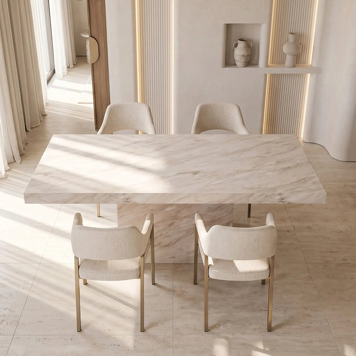 A modern dining room setup featuring a large rectangular marble-top table with a matching marble pedestal base, surrounded by four upholstered dining chairs with gold-toned legs in a bright, sunlit interior.