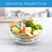Frozen vegetables in a glass bowl with text 'Defrost by Weight/Time' on a blurred kitchen background.