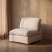 A modern off-white upholstered armless accent chair with a thick cushioned seat and backrest, set in a minimalist room with a textured beige wall, concrete floor, and sheer curtains.