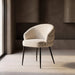 A modern upholstered accent chair with a curved back and seat, upholstered in a light beige textured fabric, supported by slim black metal legs, set in a minimalist interior with a concrete floor and textured brown walls.