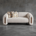 Modern beige sofa with wooden armrests and patterned cushions against a gray concrete wall.