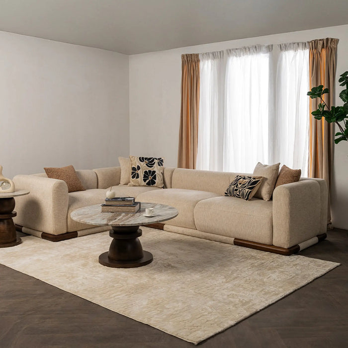 A modern beige sectional sofa in a styled living room, featuring patterned accent pillows, a round marble-top coffee table, a large cream rug, and sheer curtains with tan drapes.