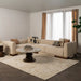 A modern beige sectional sofa in a styled living room, featuring patterned accent pillows, a round marble-top coffee table, a large cream rug, and sheer curtains with tan drapes.