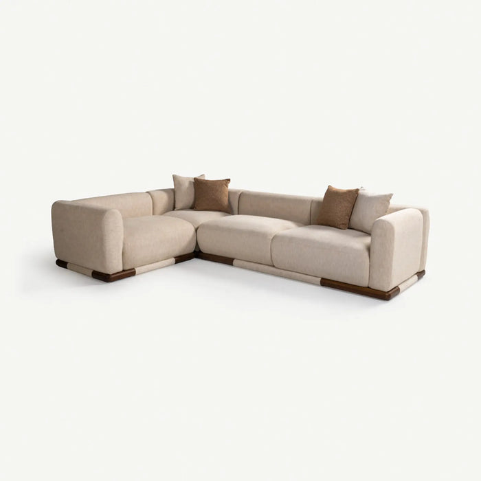 A modern L-shaped sectional sofa upholstered in light beige fabric, accented with three brown throw pillows and dark wood-look base and arm details, shown on a clean off-white background.