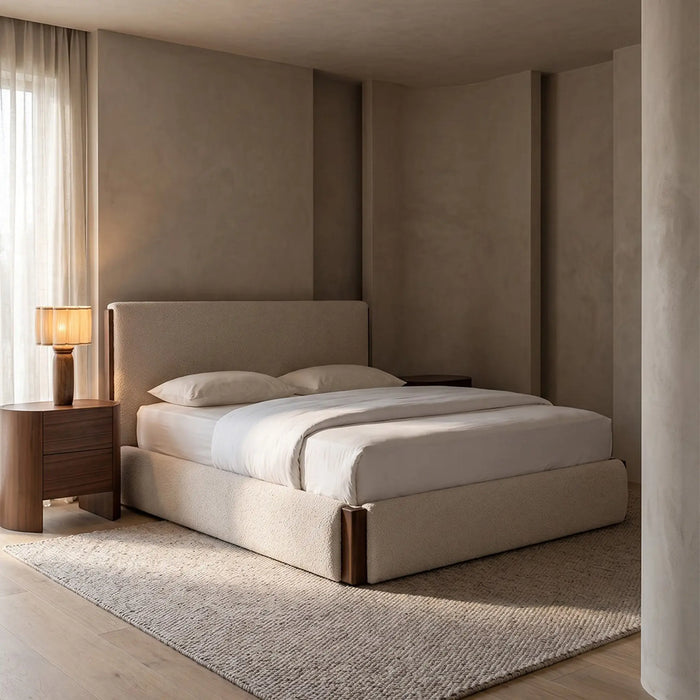 A modern upholstered bed in a minimalist bedroom with warm beige walls, a matching rug, a wooden nightstand with a lit table lamp, and light wood flooring.
